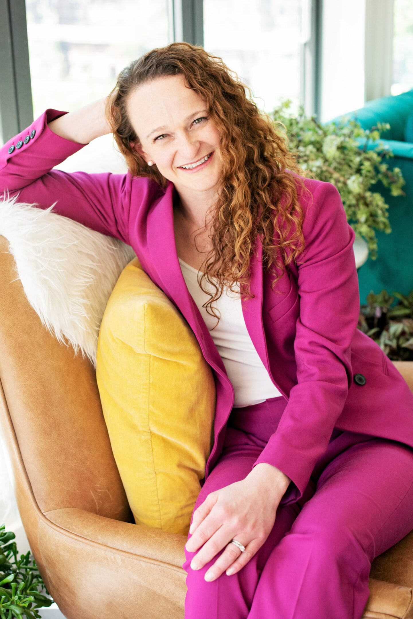 A smiling woman in a magenta suit relaxing on a couch with a yellow pillow.